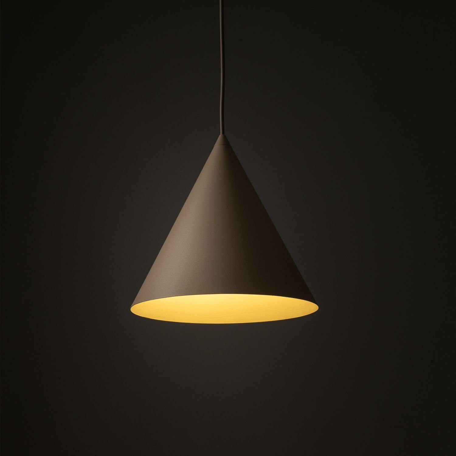 Na ciemnym tle wisi stożkowa lampa w kolorze beżowym, emitująca ciepłe, żółte światło. Lampa jest zawieszona na cienkim przewodzie.