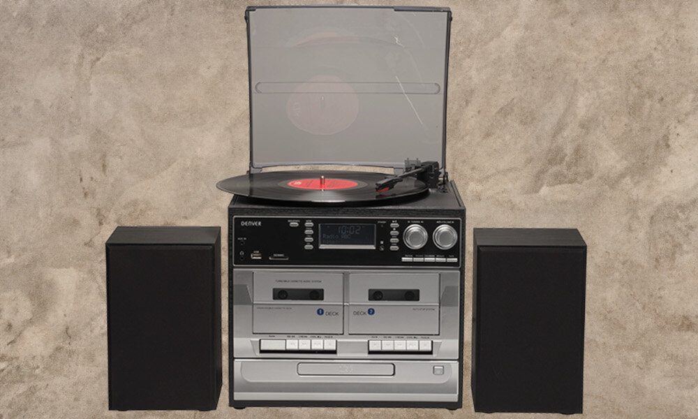 Wieża audio z gramofonem, dwoma kasetami i dwoma głośnikami. Na gramofonie znajduje się czarna płyta winylowa z czerwonym środkiem, a na froncie widoczne są przyciski i pokrętła sterujące.