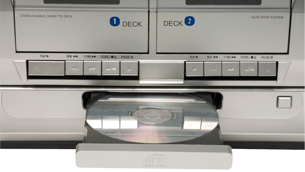 Obraz przedstawia srebrną wieżę stereo z widocznymi dwoma kasetowymi deckami oznaczonymi jako \'1 DECK\' i \'2 DECK\'. Poniżej znajduje się otwarta szuflada na płyty CD z widoczną płytą.