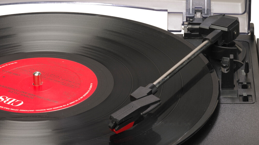 Na zdjęciu widoczny jest gramofon z czarną płytą winylową i czerwoną etykietą na środku. Ramię gramofonu jest opuszczone na płytę.