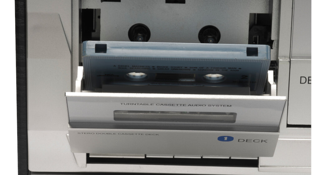 Zdjęcie przedstawia otwarty odtwarzacz kasetowy z widoczną kasetą w środku. Na urządzeniu widoczny jest napis \'TURNTABLE CASSETTE AUDIO SYSTEM\' oraz \'STEREO DOUBLE CASSETTE DECK\'.