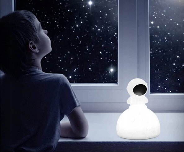 Na parapecie okna stoi lampka nocna w kształcie astronauty, emitująca delikatne światło. W tle widoczne jest rozgwieżdżone niebo.