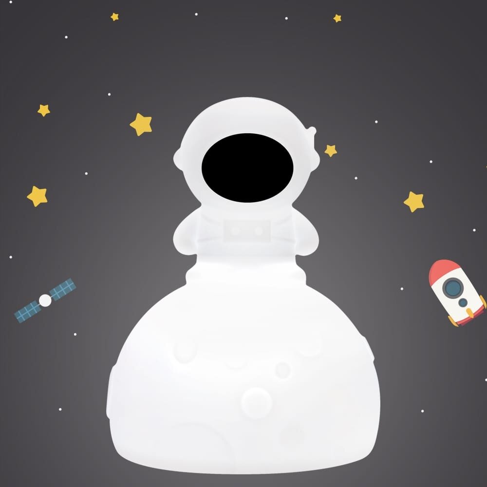 Na obrazie widoczna jest biała lampka nocna w kształcie astronauty na tle ciemnego nieba z gwiazdami, satelitą i rakietą. Astronauta ma czarną szybkę hełmu i stoi na półkolistej podstawie.