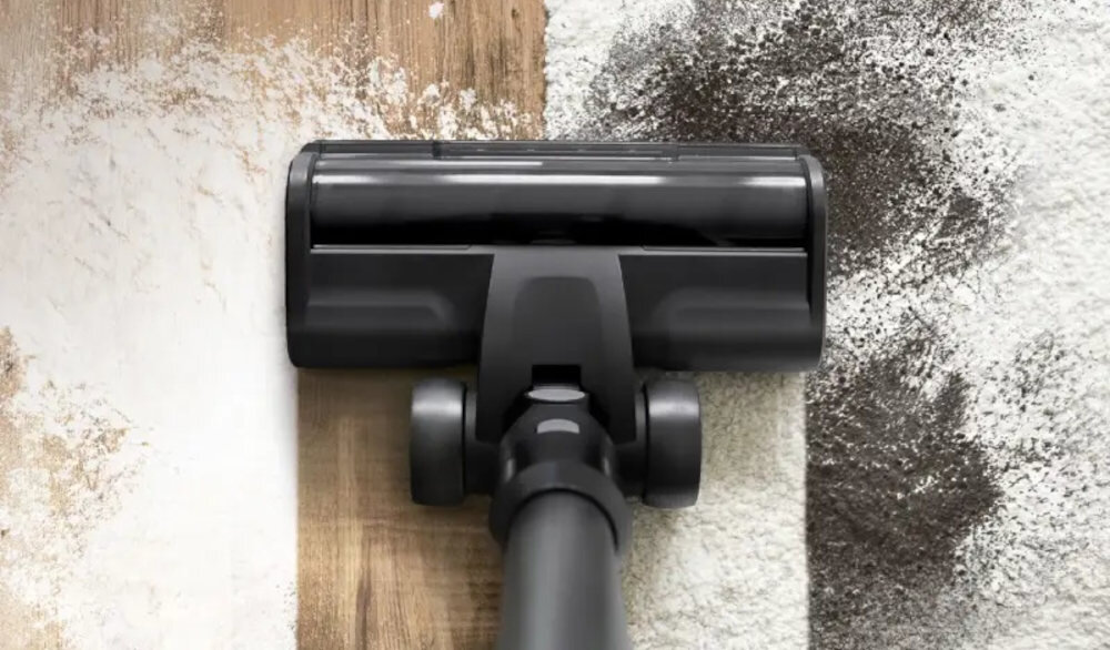 Czarna głowica odkurzacza przesuwa się po powierzchni, czyszcząc brud z podłogi. Po lewej stronie powierzchnia jest czysta, a po prawej widać zabrudzenia.