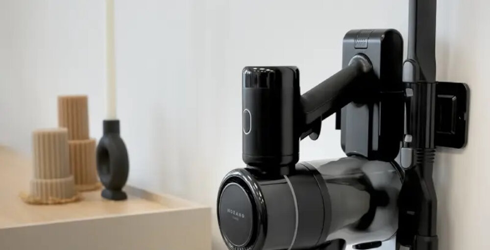 Na obrazie widoczny jest czarny odkurzacz MOZANO Handy Pro zamontowany na ścianie. W tle znajdują się dekoracyjne przedmioty na półce.