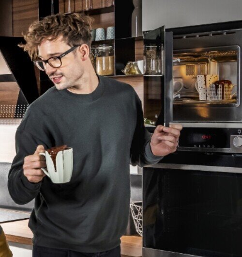 Osoba trzyma kubek z gorącą czekoladą, stojąc obok otwartej kuchenki mikrofalowej z widocznymi kubkami w środku. Na panelu kuchenki wyświetla się czas 18:41.