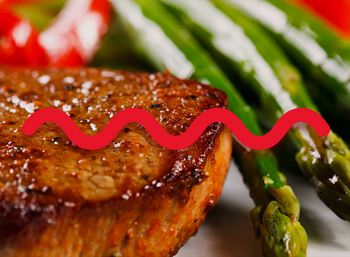 Na obrazie widoczny jest kawałek grillowanego mięsa z przyprawami, obok którego znajdują się zielone szparagi i czerwone warzywa. Przez środek obrazu przebiega czerwona, falista linia.