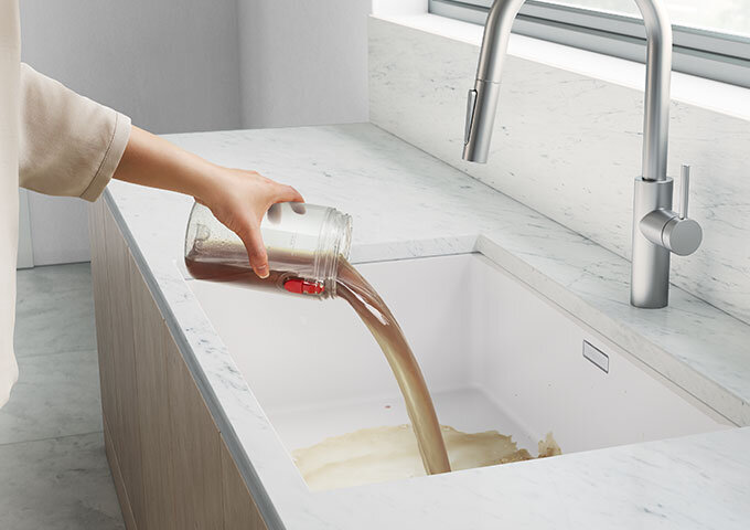 Osoba wylewa brązowy płyn z przezroczystego słoika do białego zlewu w nowoczesnej kuchni. W tle widoczne są marmurowe blaty i metalowa bateria kuchenna.