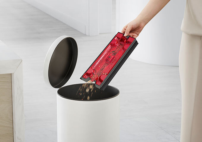 Osoba opróżnia pojemnik z czerwonym elementem do białego kosza na śmieci. W tle widoczne są jasne ściany i podłoga.
