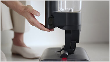 Osoba obsługuje czarny, bezprzewodowy mop elektryczny z przezroczystym pojemnikiem na wodę. W tle widoczne są jasne elementy wnętrza.