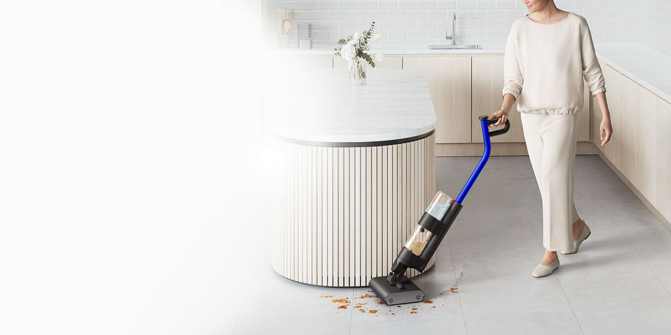 Osoba używa elektrycznego mopa z niebieskim uchwytem na jasnej podłodze w nowoczesnej kuchni. Mop zbiera rozlane płyny i resztki jedzenia obok wyspy kuchennej.