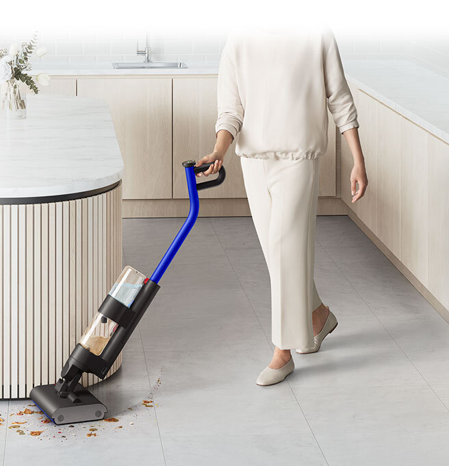 Osoba używa elektrycznego mopa z niebieską rączką do sprzątania podłogi w nowoczesnej kuchni. Na podłodze widoczne są rozsypane okruchy, które mop zbiera.