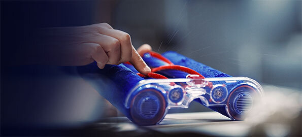 Osoba trzyma niebieski element urządzenia z przezroczystymi częściami i czerwonymi akcentami. W tle widoczne jest rozmyte otoczenie.