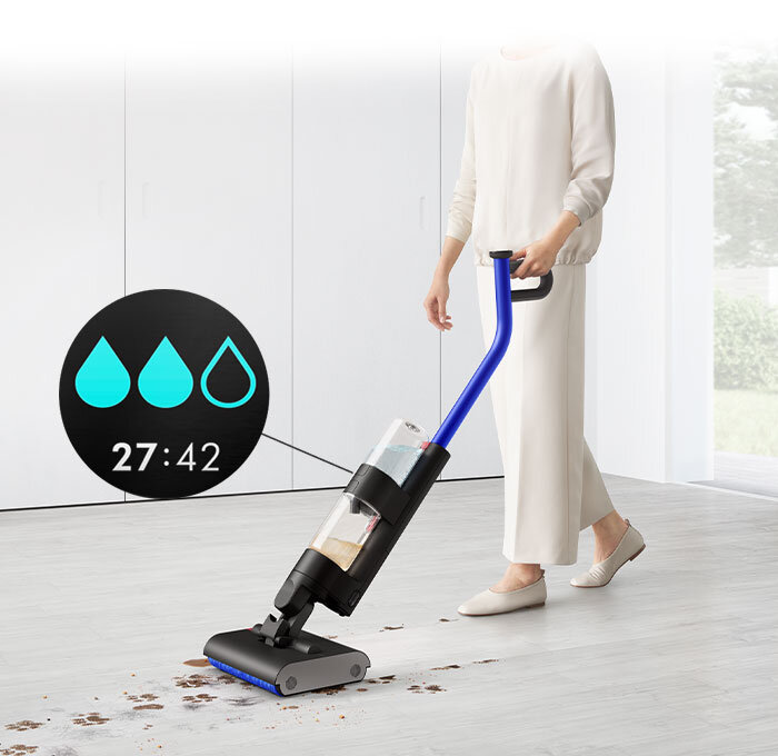 Osoba używa elektrycznego mopa z niebieskim uchwytem do czyszczenia podłogi z rozsypanymi resztkami. Na obrazie widoczny jest także okrągły wskaźnik z trzema ikonami kropli wody i czasem 27:42.