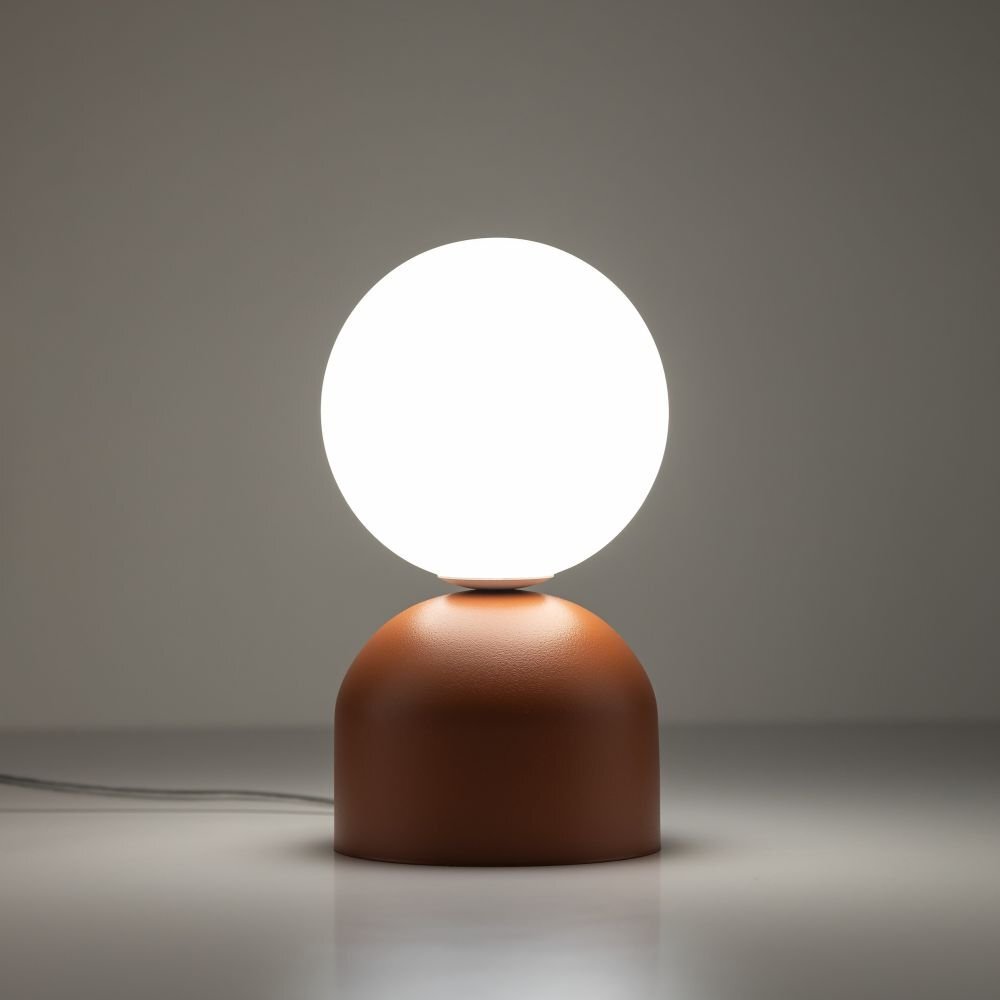 Na obrazie widoczna jest lampa stołowa z okrągłym, białym kloszem i ceglastą podstawą. Lampa stoi na gładkiej powierzchni, a w tle jest szare tło.