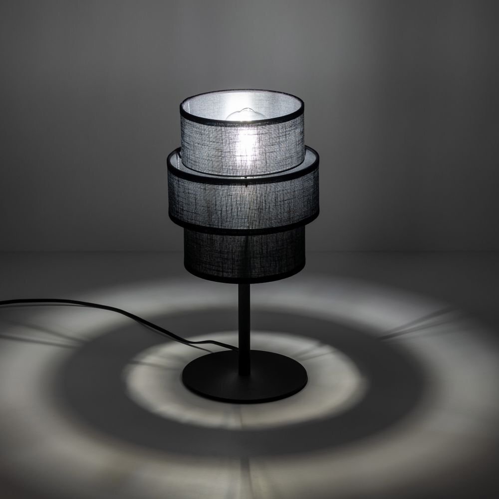 Czarna lampa stołowa z trzema cylindrycznymi abażurami, emitująca światło tworzące koncentryczne kręgi na powierzchni. Lampa stoi na okrągłej podstawie z widocznym przewodem zasilającym.