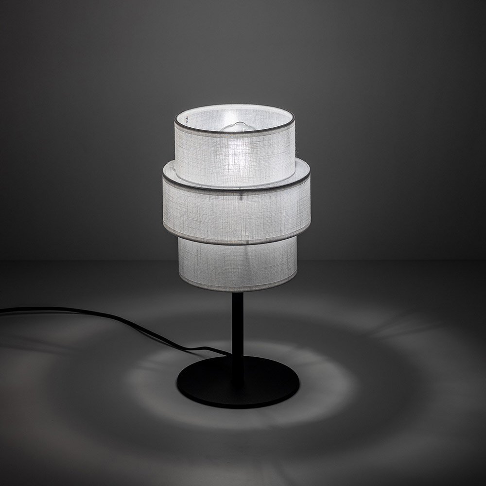 Na obrazie widoczna jest lampa stołowa z białym abażurem o cylindrycznym kształcie, umieszczona na czarnej, metalowej podstawie. Lampa emituje delikatne światło, tworząc wzory na powierzchni, na której stoi.