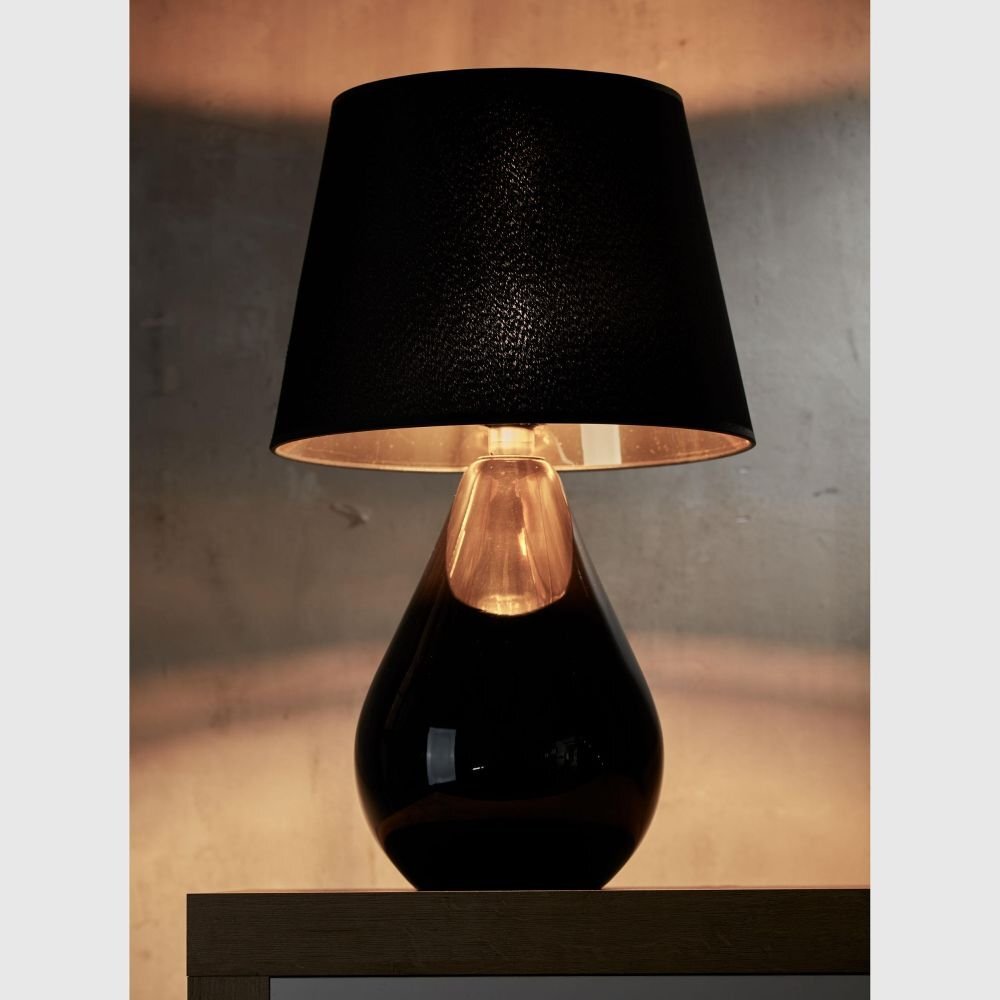 Czarno-złota lampa stołowa z abażurem w kształcie stożka, umieszczona na drewnianej powierzchni. Światło lampy tworzy ciepłą poświatę na ścianie w tle.