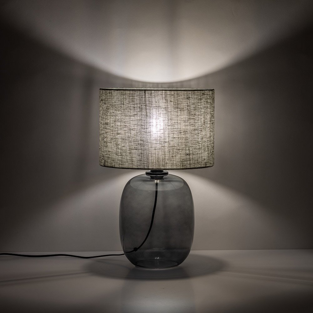 Lampa stołowa z przezroczystą, szklaną podstawą i cylindrycznym abażurem w kolorze grafitowo-zielonym. Światło rozprasza się na ścianie, tworząc efektowne wzory.