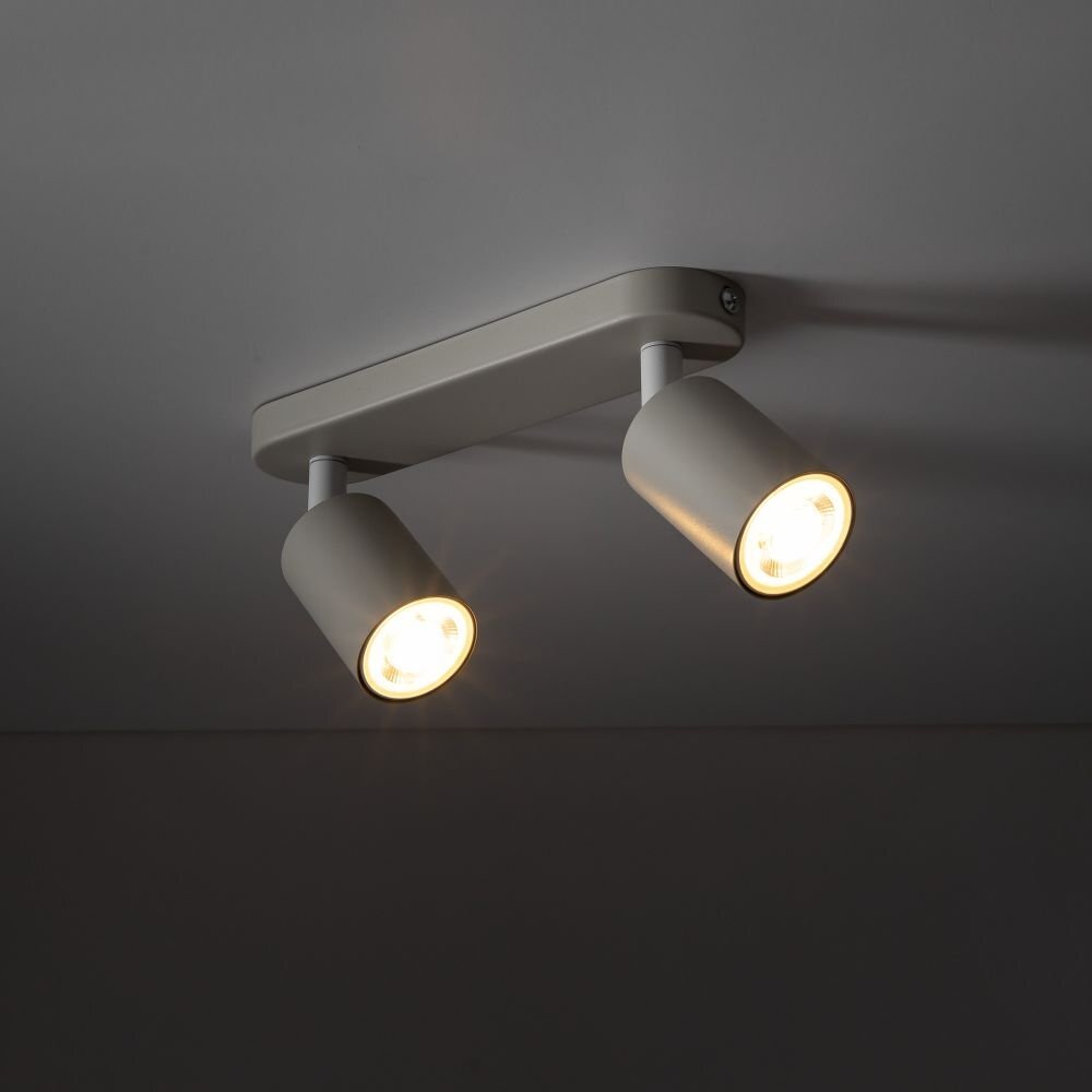 Na obrazie widoczna jest beżowa lampa sufitowa z dwoma regulowanymi reflektorami, zamontowana na suficie. Reflektory emitują ciepłe światło.