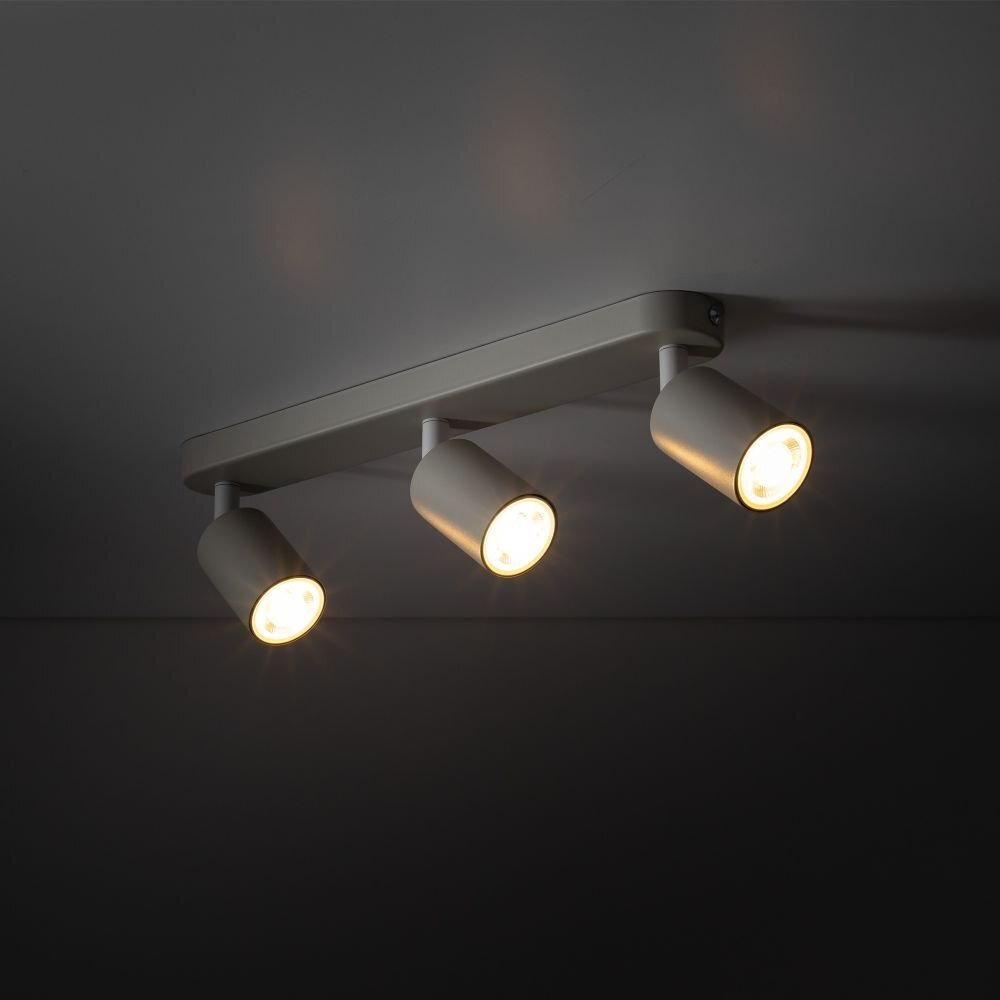 Na obrazie widoczna jest szara lampa sufitowa z trzema regulowanymi reflektorami, emitującymi ciepłe światło. Lampa zamontowana jest na ciemnym suficie.