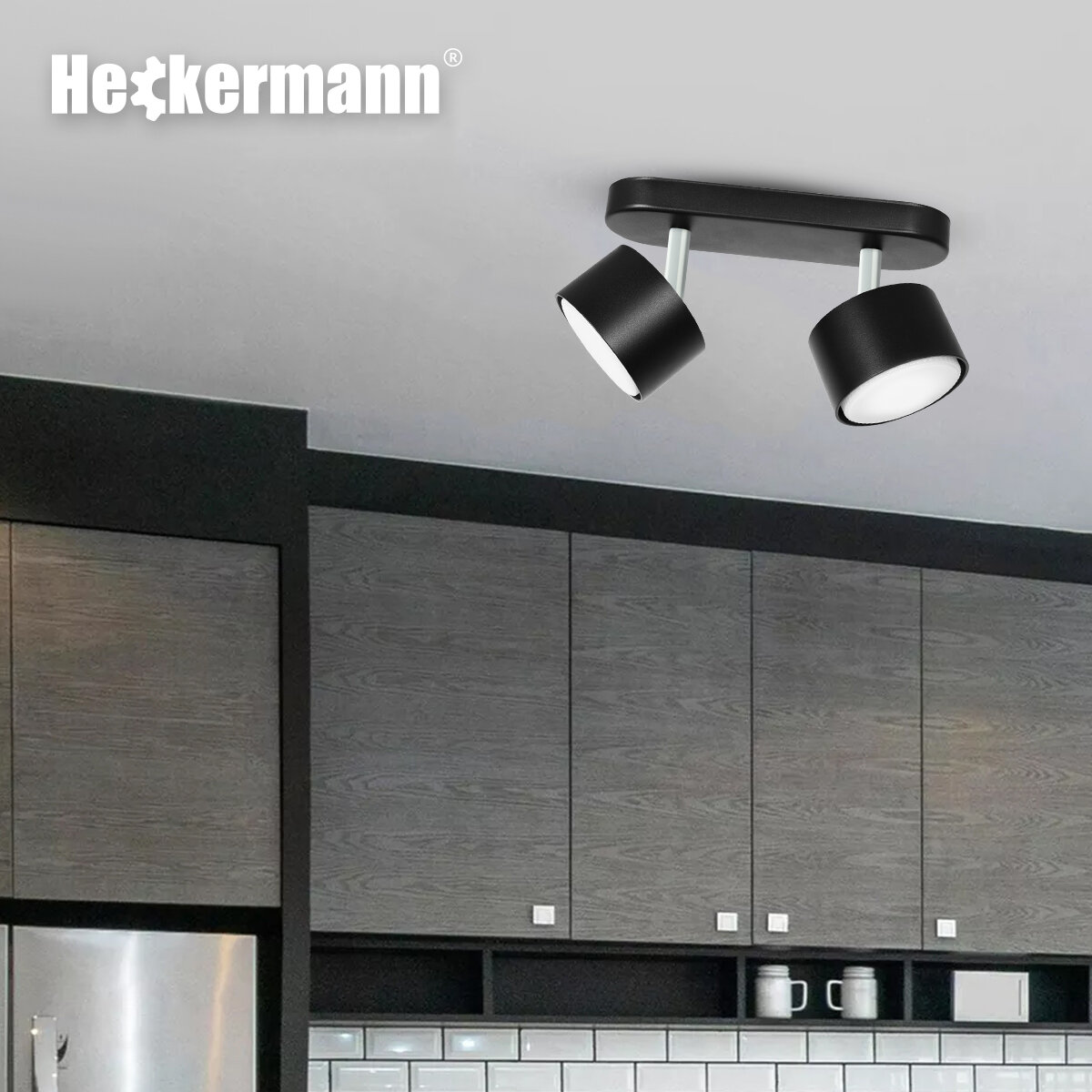 Na obrazie widoczna jest czarna lampa punktowa z dwoma głowicami zamontowana na suficie. W tle znajdują się szafki kuchenne oraz logo \'Heckermann\'.