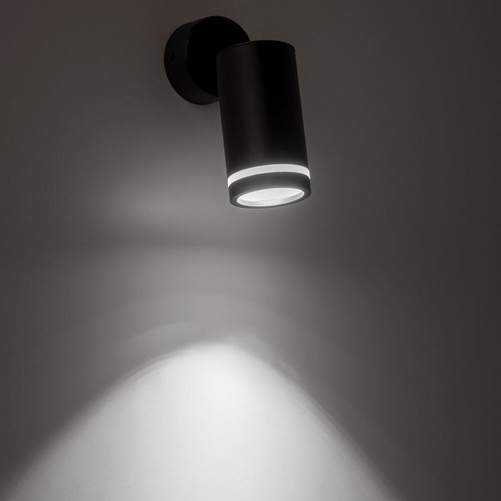 Czarny reflektor sufitowy z metalowym korpusem emituje światło skierowane w dół, tworząc jasną plamę na ścianie. Reflektor ma cylindryczny kształt i jest zamontowany na okrągłej podstawie.