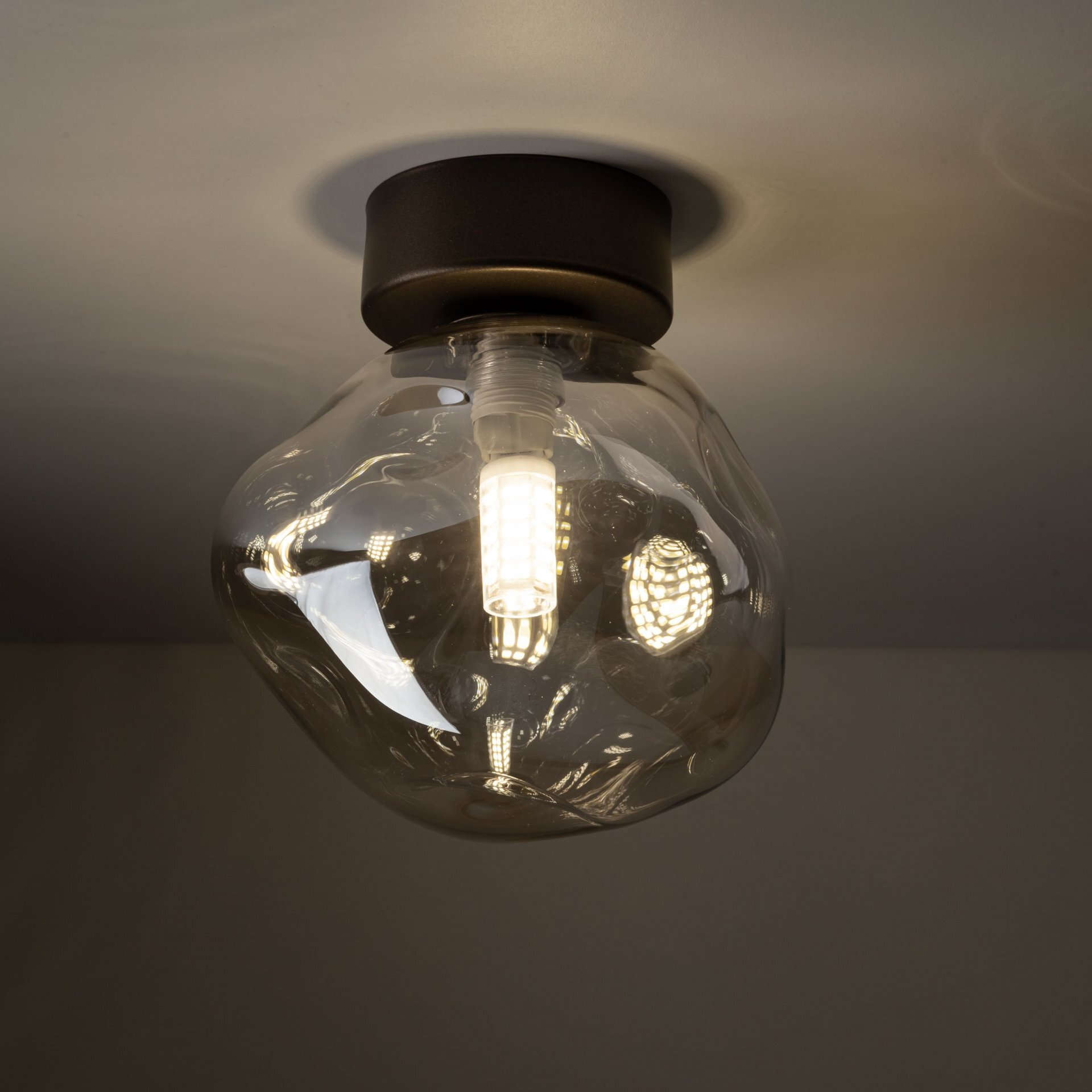 Lampa sufitowa z bursztynowego szkła o nieregularnym kształcie, z widoczną żarówką LED wewnątrz. Montowana jest na ciemnej, okrągłej podstawie przylegającej do sufitu.