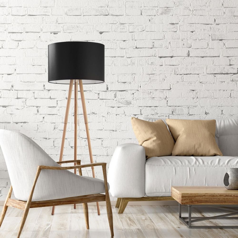 Wnętrze z białą ceglaną ścianą, na której tle stoi czarna lampa podłogowa z drewnianymi nogami. Obok znajduje się biały fotel i sofa z beżowymi poduszkami oraz drewniany stolik kawowy.