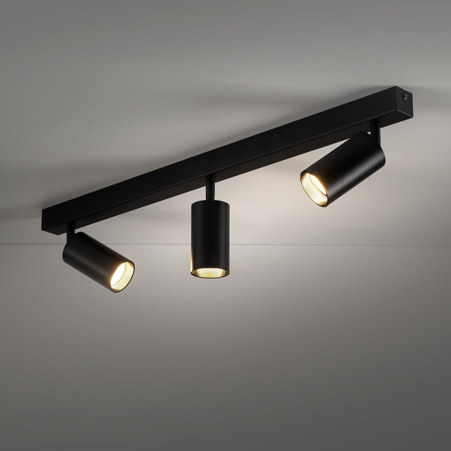 Czarna lampa sufitowa z trzema regulowanymi reflektorami w kształcie cylindrów, zamontowana na prostokątnej listwie. Reflektory emitują ciepłe światło, a wnętrza są złote.