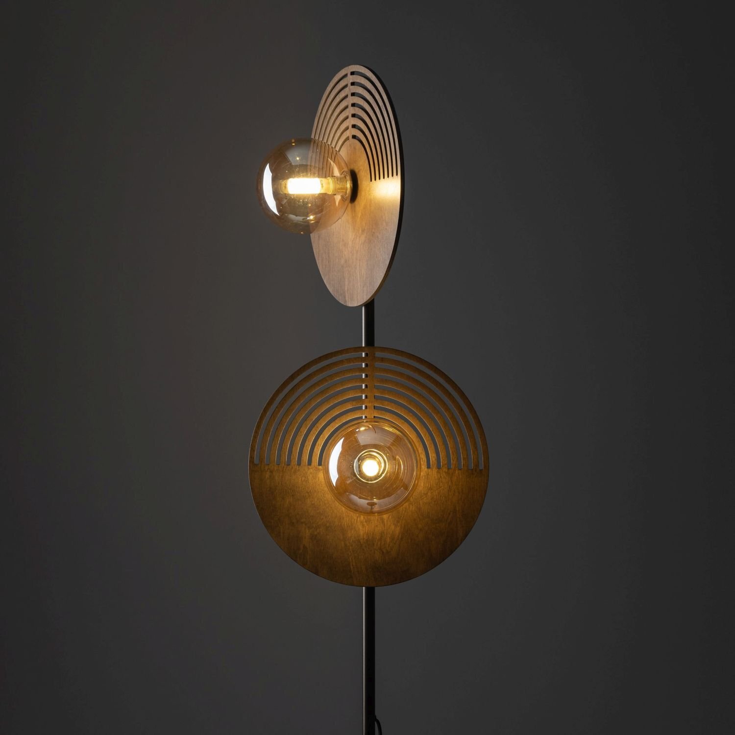 Lampa podłogowa z dwoma okrągłymi kloszami, każdy z żarówką, umieszczona na ciemnym tle. Klosze mają drewniane elementy z wycięciami w kształcie koncentrycznych okręgów.