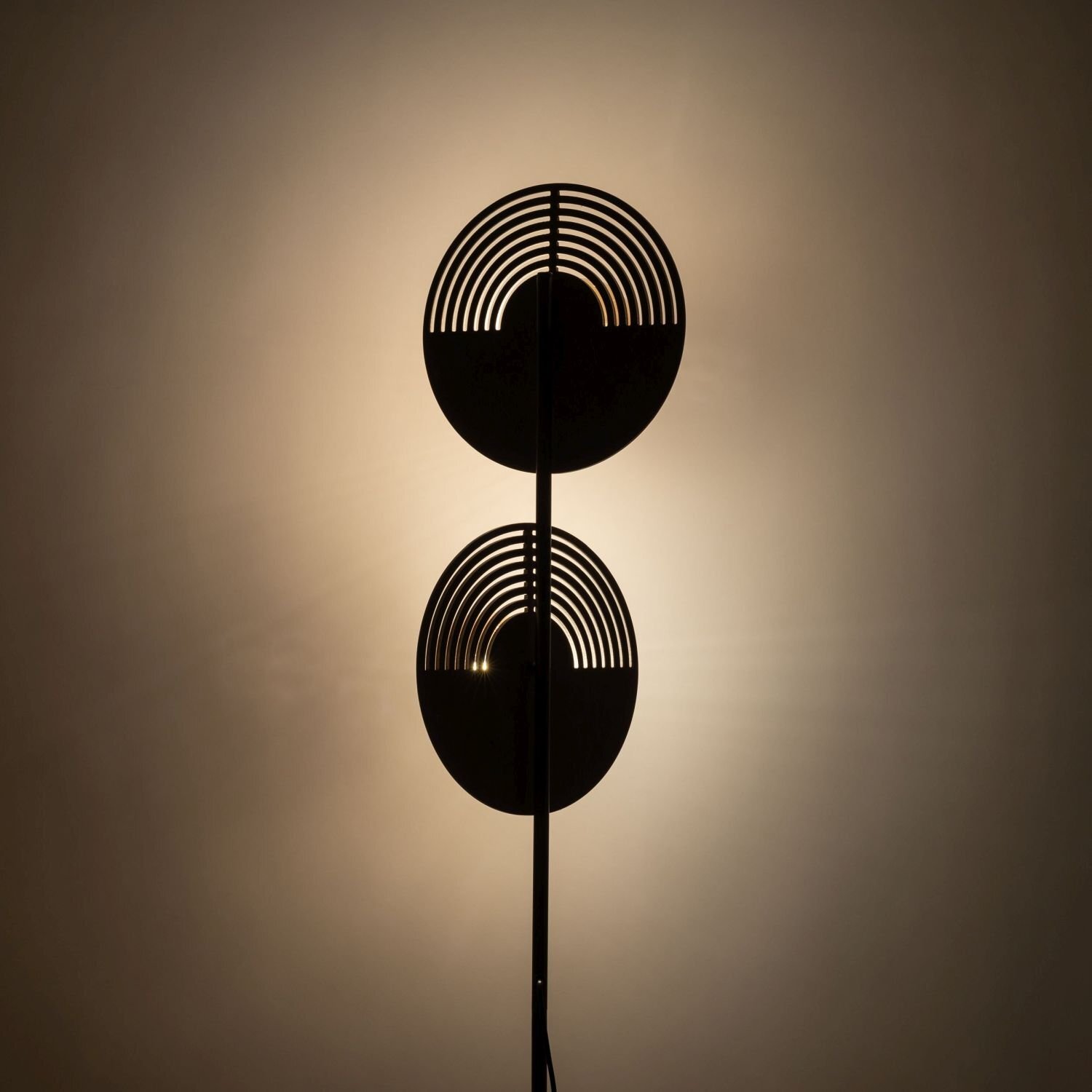 Lampa podłogowa z dwoma okrągłymi kloszami o geometrycznym wzorze, emitująca ciepłe światło na ścianę. Klosze mają ciemny kolor i są umieszczone na pionowym stelażu.