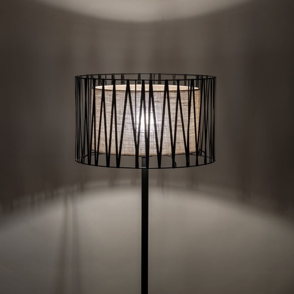Lampa podłogowa z czarnym stelażem i abażurem z juty, emitująca ciepłe światło. Konstrukcja abażuru składa się z pionowych, czarnych prętów otaczających materiałowy klosz.