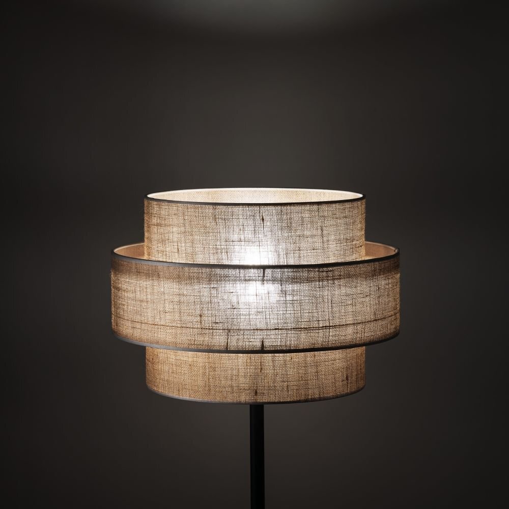 Lampa podłogowa z czarnym stelażem i jutowym abażurem o strukturze tkaniny. Abażur ma cylindryczny kształt z widocznymi warstwami, przez które prześwituje światło.