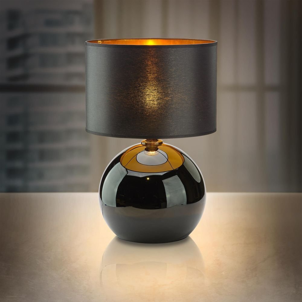 Czarna lampa stołowa z kulistą podstawą i cylindrycznym abażurem, wewnątrz złotym. Lampa stoi na gładkiej powierzchni, a w tle widać rozmyte okno.