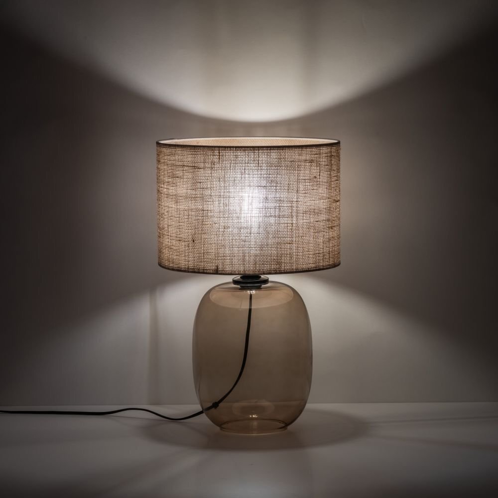 Na obrazie widoczna jest lampa stołowa z bursztynową, szklaną podstawą i jutowym abażurem. Lampa emituje ciepłe światło, tworząc przytulną atmosferę.