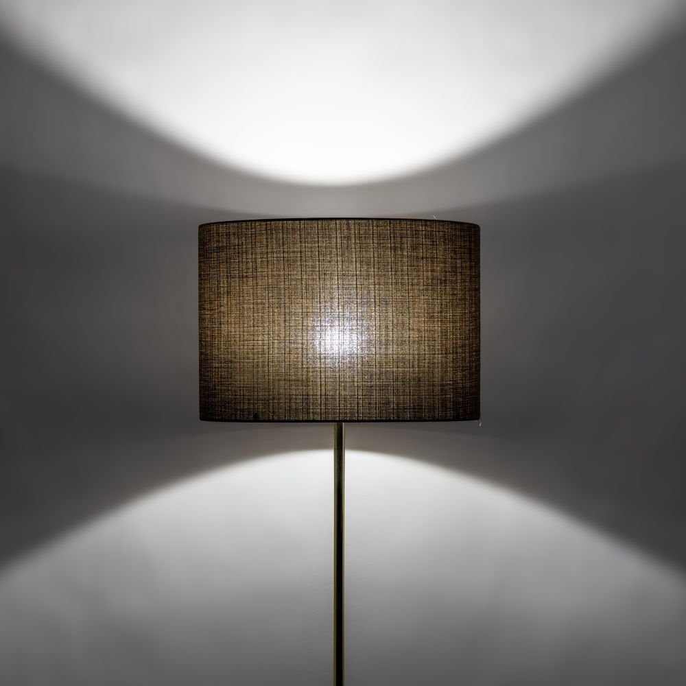 Lampa podłogowa z ciemnym abażurem, emitująca światło tworzące wzór na ścianie. Podstawa lampy jest smukła i metaliczna.