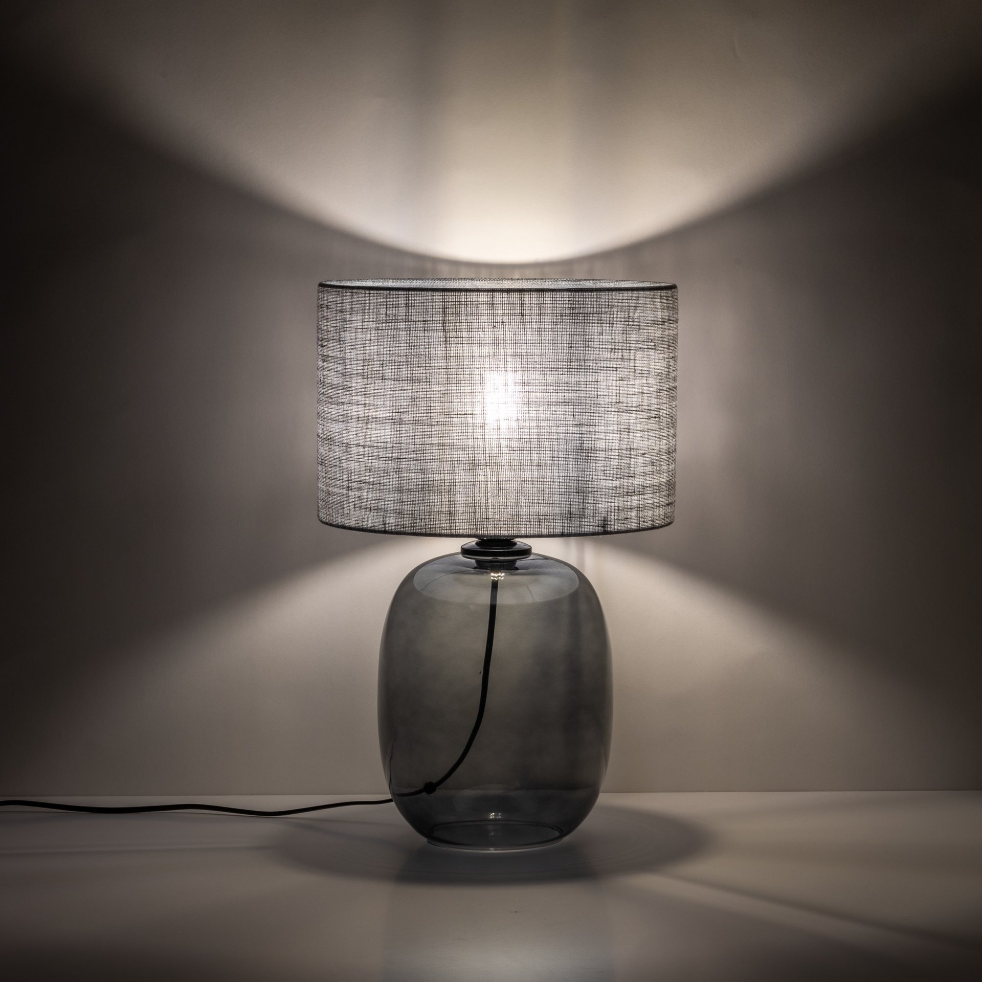 Na obrazie widoczna jest nowoczesna lampa stołowa z grafitowym abażurem i szklanym, przezroczystym korpusem. Lampa emituje miękkie światło, tworząc delikatne cienie na ścianie.