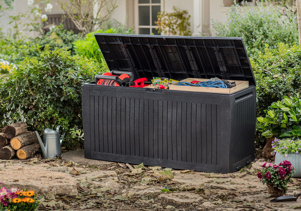 Otwarta skrzynia ogrodowa w kolorze antracytowym, wypełniona narzędziami i akcesoriami, stoi na tle zieleni ogrodowej. Obok skrzyni znajduje się konewka i stos drewna.