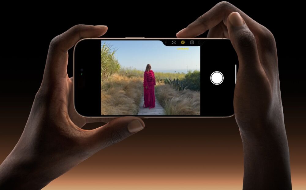 Osoba trzyma smartfon, na ekranie którego widać zdjęcie kobiety w czerwonej sukni stojącej na ścieżce wśród traw. Interfejs aparatu pokazuje przycisk migawki i kilka ikon na górze ekranu.