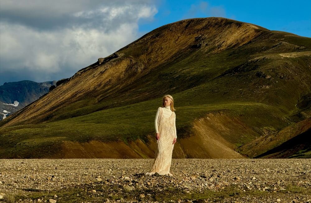 Osoba w jasnej sukni stoi na kamienistym terenie z dużym wzgórzem w tle. Niebo jest częściowo zachmurzone.