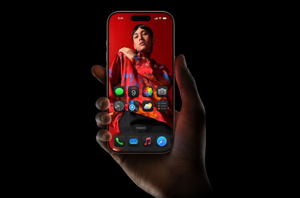 Osoba trzyma smartfon z wyświetlaczem pokazującym kolorowy ekran główny z ikonami aplikacji. W tle ekranu widoczna jest postać ubrana w kolorową odzież na czerwonym tle.