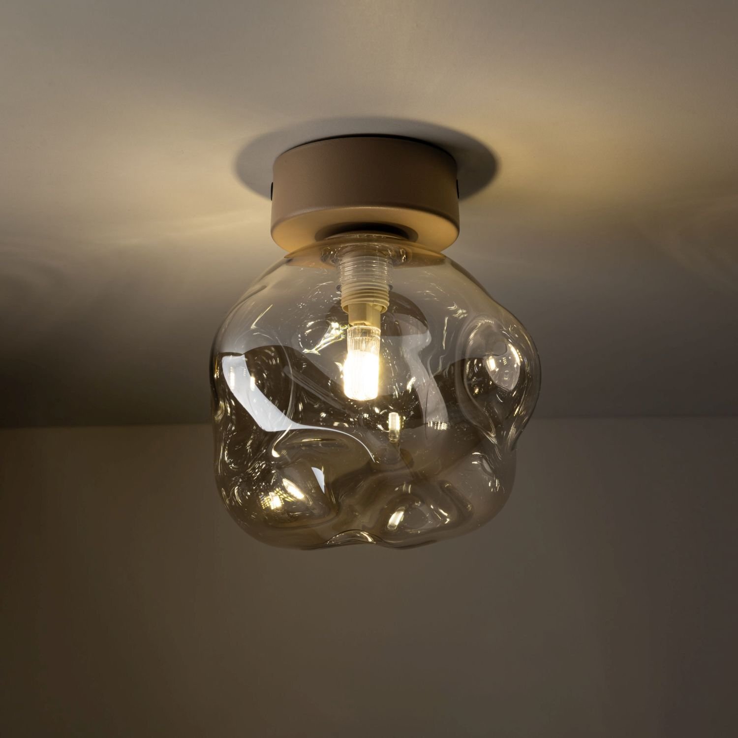 Na obrazie widoczna jest lampa sufitowa z przezroczystym, szklanym kloszem o nieregularnym kształcie. Wewnątrz klosza znajduje się żarówka emitująca ciepłe światło.