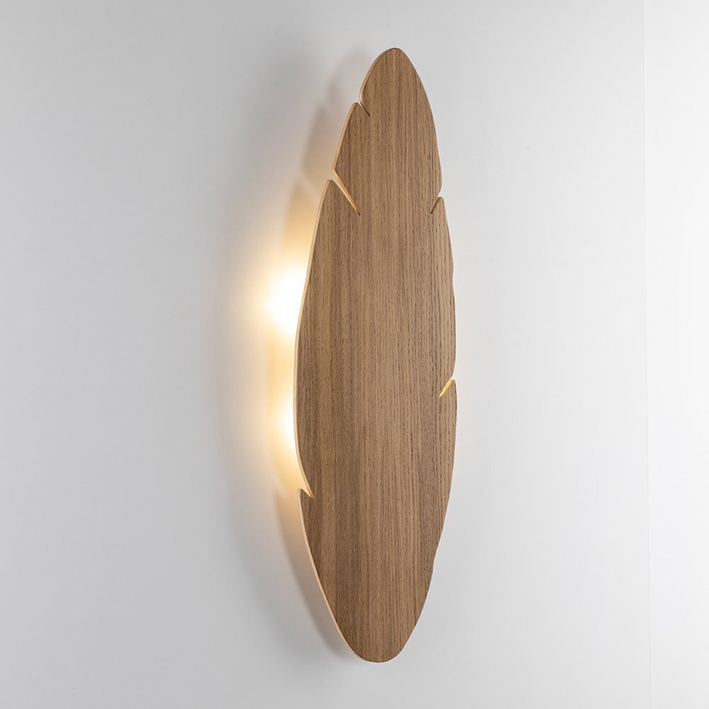 Na obrazie widoczny jest kinkiet w kształcie liścia, wykonany z drewna, z delikatnym podświetleniem od tyłu. Kinkiet zamontowany jest na jasnej ścianie.