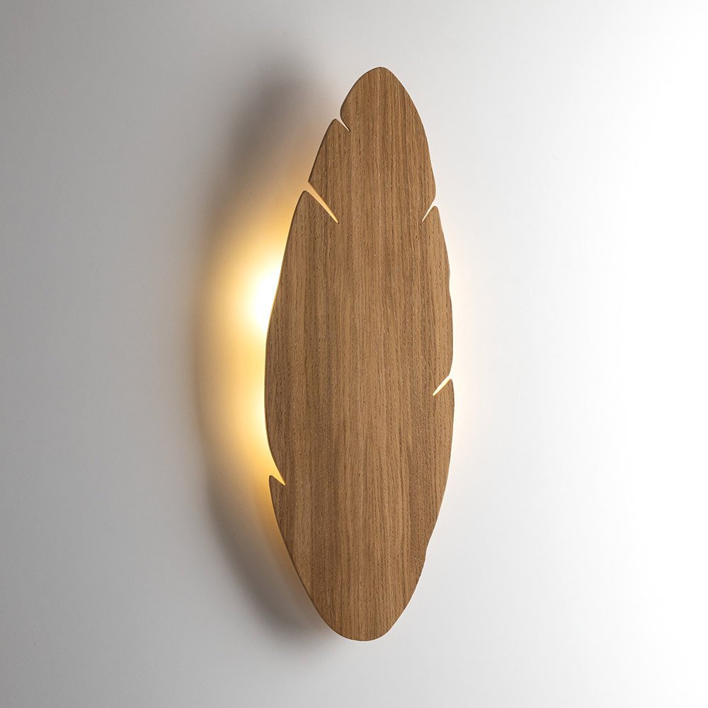 Na obrazie widoczny jest kinkiet w kształcie liścia, wykonany z drewna, z delikatnym podświetleniem od tyłu. Lampa ma nowoczesny design i jest zamontowana na jasnej ścianie.