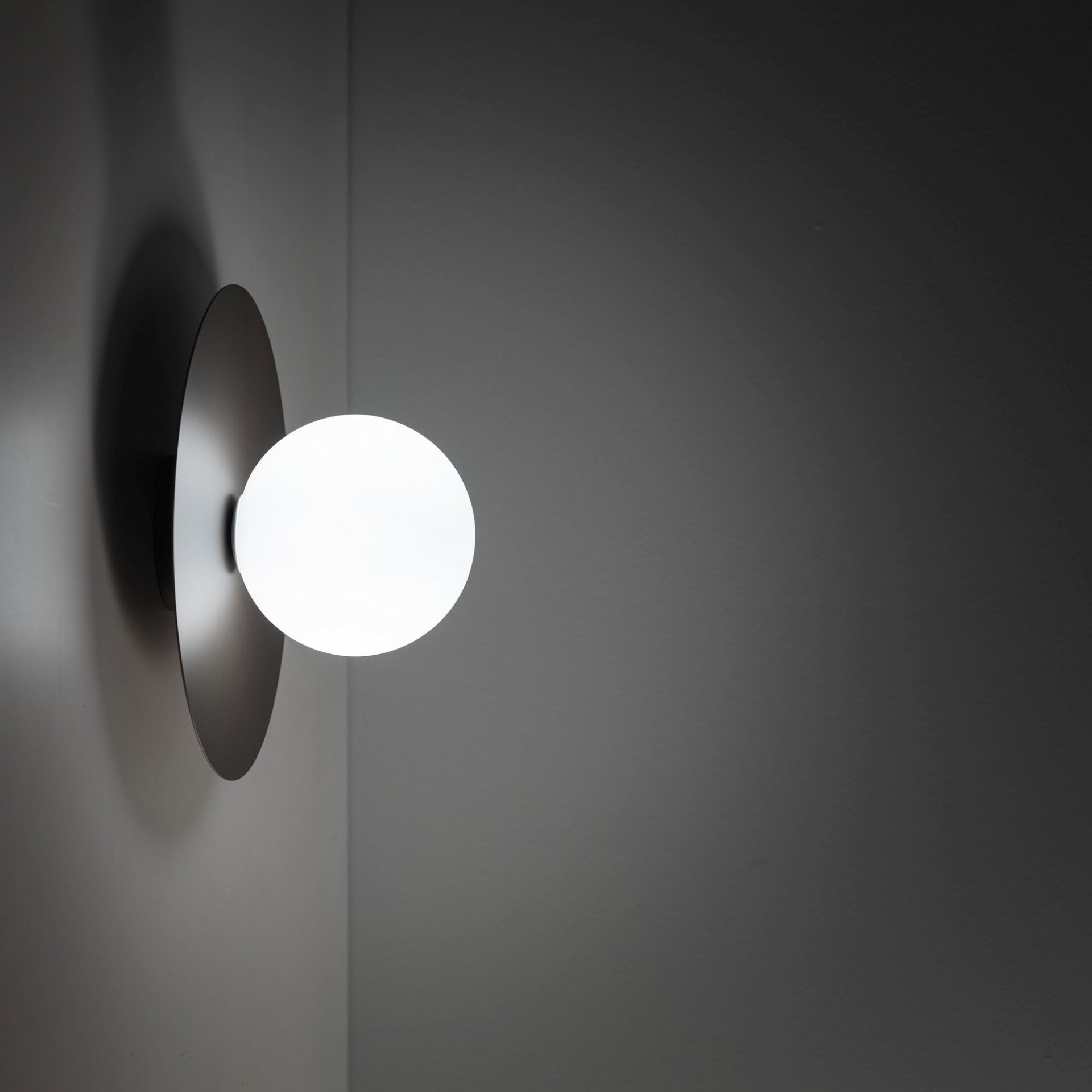 Na obrazie widoczna jest lampa ścienna z okrągłym, białym kloszem zamontowanym na ciemnym, okrągłym uchwycie. Światło lampy oświetla ścianę, tworząc delikatny efekt świetlny.