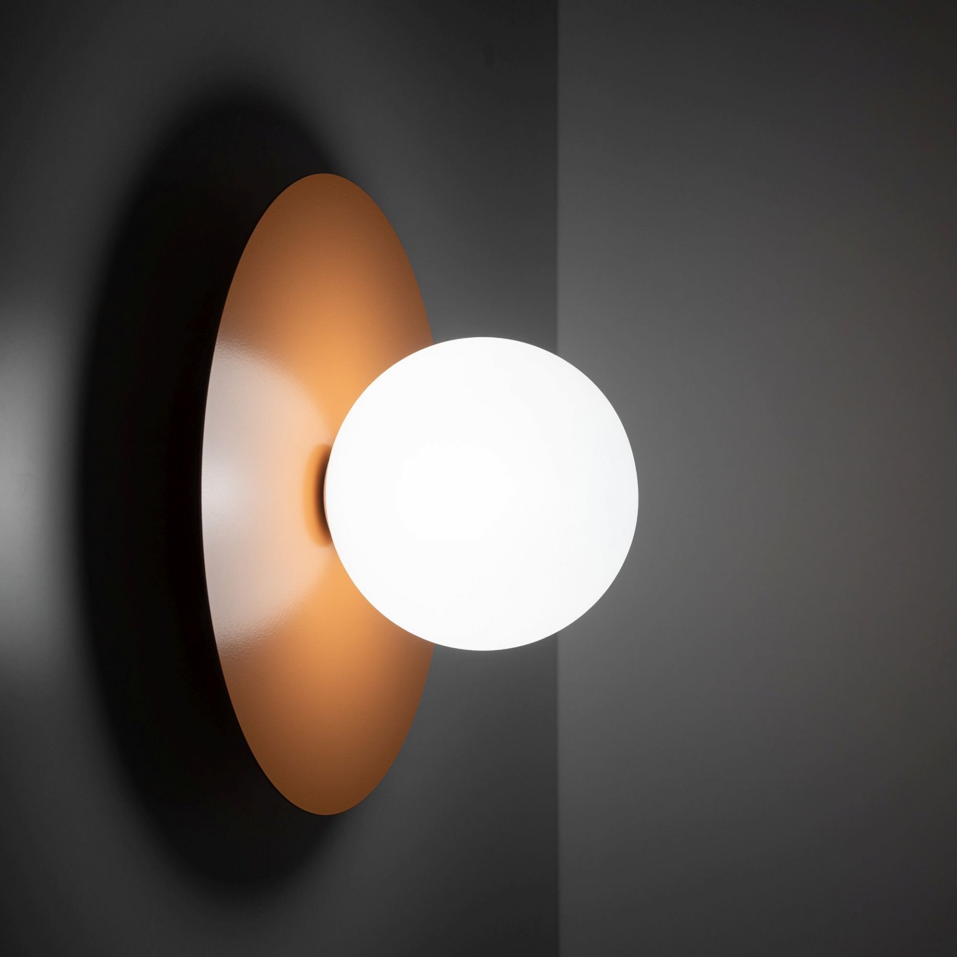 Na obrazie widoczna jest nowoczesna lampa ścienno-sufitowa z okrągłym, białym kloszem i pomarańczowym, metalowym dyskiem. Lampa jest zamontowana na ciemnej ścianie.