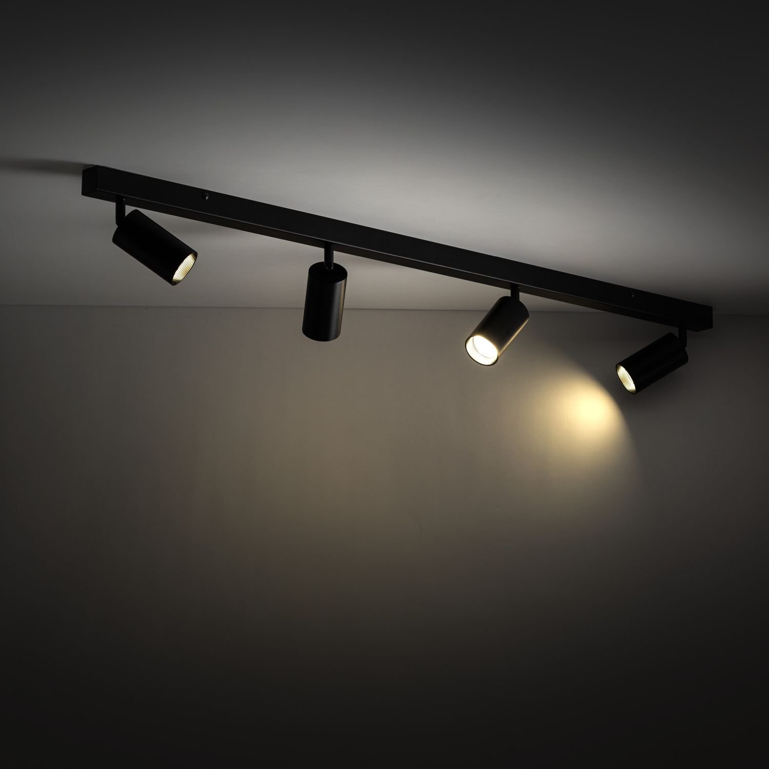Czarna lampa sufitowa z czterema regulowanymi reflektorami, z których dwa są włączone, emitując ciepłe światło na ścianę. Lampa jest zamontowana na suficie i wykonana z metalu.