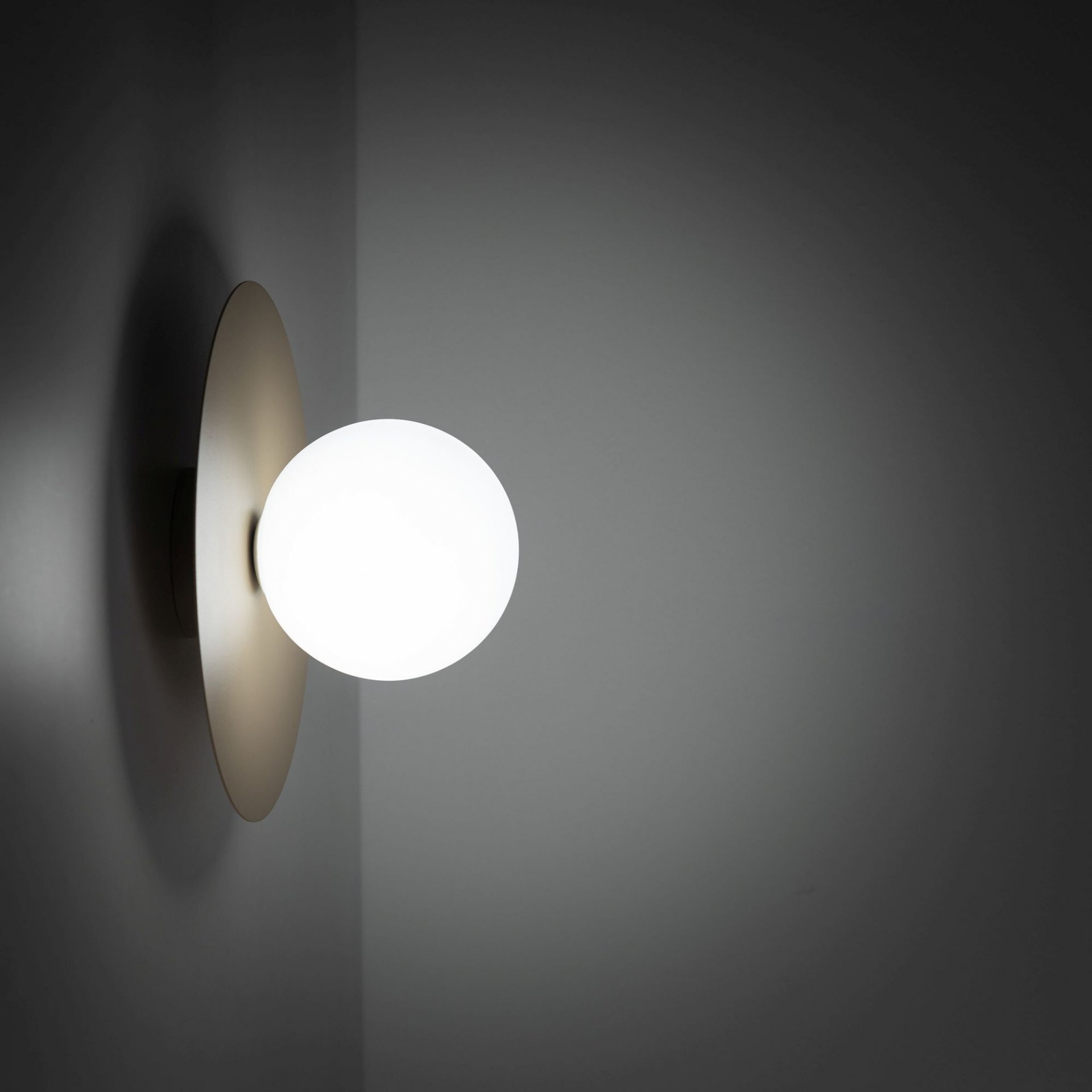 Na obrazie widoczna jest nowoczesna lampa ścienno-sufitowa z okrągłym, białym kloszem i beżową podstawą. Lampa emituje jasne światło, tworząc delikatny efekt na ścianie.