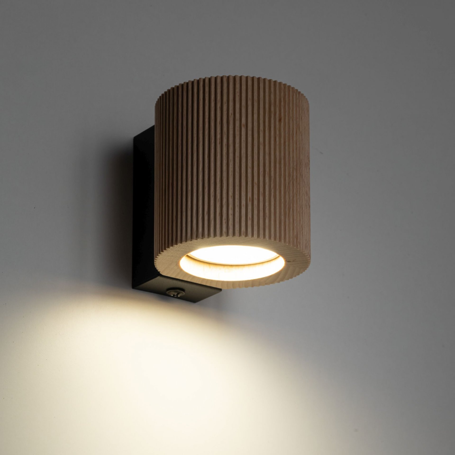 Na obrazie widoczna jest lampa ścienna z cylindrycznym kloszem wykonanym z drewna o żłobionej powierzchni. Lampa emituje ciepłe światło skierowane w dół, zamontowana na ciemnej podstawie przylegającej do ściany.